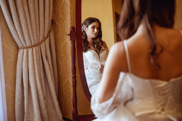 Fototapeta premium Bride in long traditional white dress standing in front of mirror in hotel room before wedding