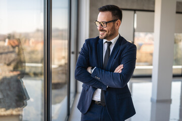 Young happy successful businessman with arms crossed smiling at corporate office