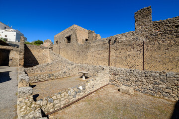 Obraz premium Ancient stone structures and weathered masonry walls stand beneath a vibrant blue sky at the House of the Lararium of Achilles in Pompeii, Italy. The open archaeological site reveals historic textures