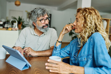 Portrait of a lovely tablet senior mature couple using a laptop together and having fun drinking coffee or tea sitting at a table at home
