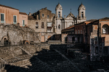 Teatro Romano E Odeon In Catania, Sicily: Ancient Roman Theater Ruins With Stone Seating, Historic Buildings, And Church Towers Under Clear Daylight