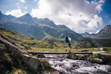 Backpacker crossing wooden bridge over mountain stream on hiking trail in Austrian Alps mountains. Solo hike adventure