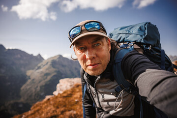 Tired hiker with backpack taking selfie on smartphone after successfully climbed mountain peak in Austrian Alps. Outdoors adventure lifestyle