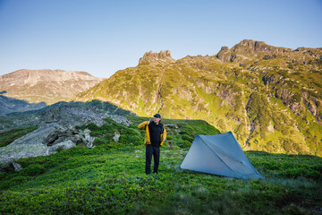 Wild camping under mountain peaks. Solo hiker standing at ultralight tent after setting campsite during trekking in wilderness