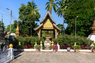 Ornate Buddhist shrine with a golden Buddha statue stands amid lush greenery and palm trees in Luang Prabang, Laos. Decorative details, potted flowers, and golden finials create a vibrant Southeast
