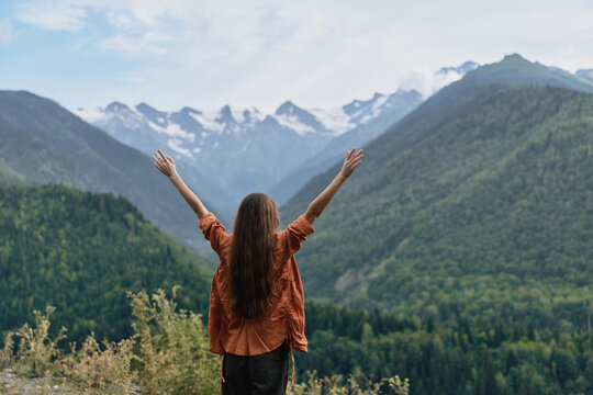 Person stands with arms raised at a scenic mountain viewpoint, enjoying a vast forested landscape and snowcapped peaks in the distance, radiating freedom and awe - Powered by Adobe