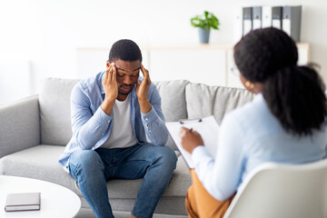 Depressed black man rubbing his temples, consulting psychologist, sitting on couch at counselor's office. Unhappy Afro guy with nervous breakdown or PTSD having session with psychiatrist at clinic