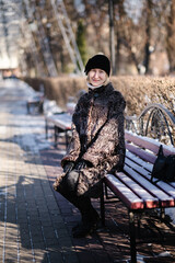 Woman sitting on a park bench in winter, wearing a warm coat and hat, smiling gently at the camera, with bare trees, snow patches, and soft daylight around.