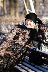 Woman relaxing on a park bench in winter, wearing a warm fur coat and black hat, smiling at the camera in bright sunlight with bare trees and snow around.