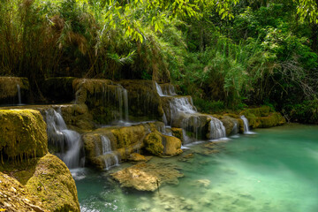 Layered limestone cascades flow into a tranquil turquoise pool surrounded by dense tropical forest at Kuang Si Falls in northern Laos. Sunlight filters through lush foliage, highlighting the natural © Florent