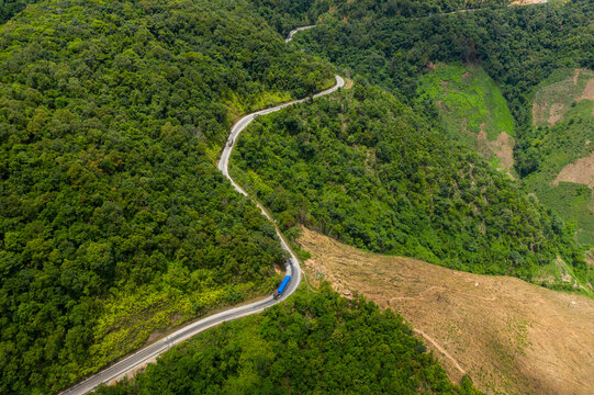 A serpentine mountain road curves through dense green forest and open hillside in the highland region between Phou Khoun and Luang Prabang, Laos. The scene captures vibrant vegetation, contrasting