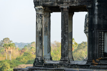 Fototapeta premium Aged sandstone pillars frame the view from the edge of Angkor Vat, Cambodia, revealing tropical foliage and soft morning light beyond. The textured surfaces and subtle carvings highlight the temple's