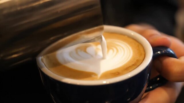 Barista pouring latte art heart in coffee cup, professional coffee making process in cafe, slow motion espresso and milk foam texture.