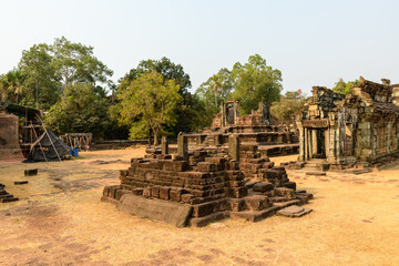 Weathered sandstone structures and stepped platforms stand amid dry grass and scattered trees at Bakong temple in Cambodia. The scene features historic architecture, subtle textures, and warm natural
