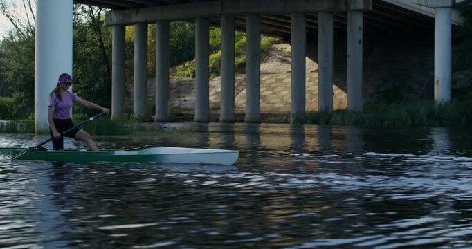 A girl in a canoe passes under a bridge during training. A man and a woman canoeist confidently paddle along the river.