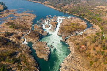 Obraz premium Expansive aerial scene of the Chutes de Khone waterfalls on the Mekong River in southern Laos. Whitewater rapids cascade through rugged rocky channels, surrounded by dry forest and a wide river plain