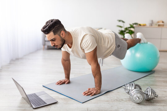 Strong Arab guy standing in plank in front of laptop, using fitness ball, working out to online sports tutorial at home. Young middle Eastern man exercising abs muscles. Domestic training concept
