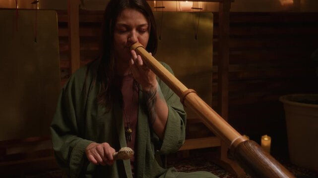 Medium shot of focused Caucasian female musician sitting in lotus posture on carpet in studio, performing while playing didgeridoo flute and rattle, during spiritual practice