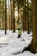 Cute young woman wearing a warm down coat standing in winter forest, looking aside. Sun rays shine through snowy trees. Mental health, calm walk, winter nature, cozy lifestyle.