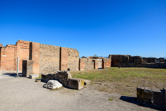 Ancient stone and brick walls of the Comitium stand in an open archaeological area of Pompeii, Italy. Bright sunlight reveals textured masonry and scattered fragments against a vivid blue sky.