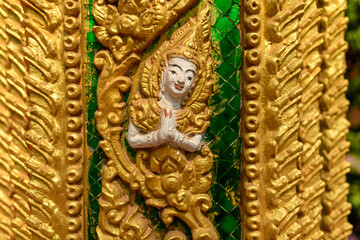 Obraz premium Close-up of an ornate gold-painted relief depicting a traditional deity figure with hands in a prayer gesture, set against a vibrant green glass mosaic at Vat Simuong temple in Vientiane. Intricate