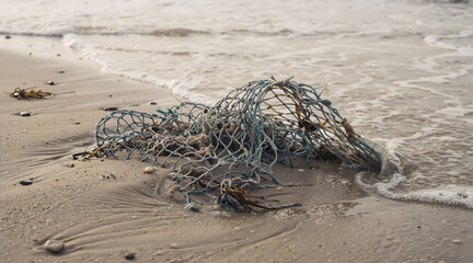 Obraz premium Discarded blue fishing net on a sandy beach. Ocean pollution and marine debris concept. Plastic waste and seaweed tangled on the shoreline. Environmental ecology and health impact