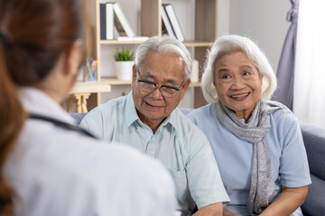 Happy retired senior asian couple sitting on sofa listening to medical advice from female doctor with joyful smile and feeling relieved about healthy life at home