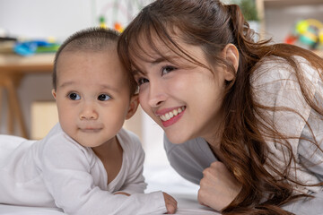 Happy asian mother lying next to adorable baby son smiling cheerfully on comfortable bed in cozy bedroom having fun together with love care and warm bond
