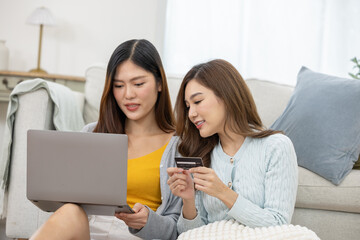 Excited asian couple women shopping online with credit card laptop computer sitting in comfortable living room feeling happy purchasing ecommerce product internet payment finance banking lifestyle