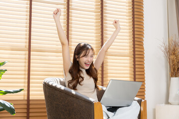 Excited young asian woman sitting armchair raising arms up celebrating success victory feeling happy with laptop computer working at home living room near window blind