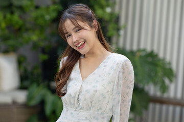 Happy asian woman standing outdoors with cheerful smile looking at camera expressing joy wearing white fashion dress with floral pattern in summer garden with greenery background