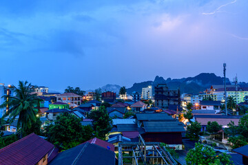 A bolt of lightning streaks across the dusk sky above the rooftops of Vang Vieng, Laos, with illuminated houses and palm trees in the foreground. Dramatic limestone mountains form a dark silhouette in