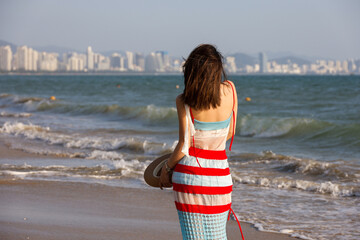 Young slim woman in summer dress standing on a beach on sea waves and city background. Summer travel and vacation