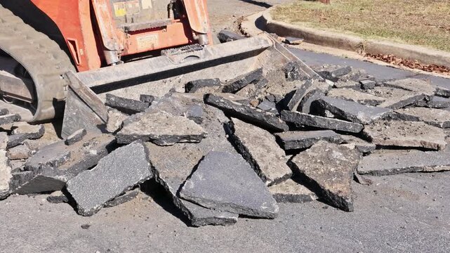 Construction worker uses skid steer loader to break up old asphalt in work area.