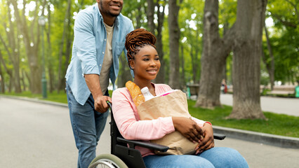 Millennial black handicapped woman in wheelchair holding paper bag with food, going shopping with...