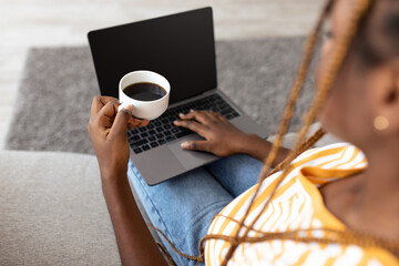 Over shoulder shot of young black woman with stylish hair sitting on couch at home, using laptop,...