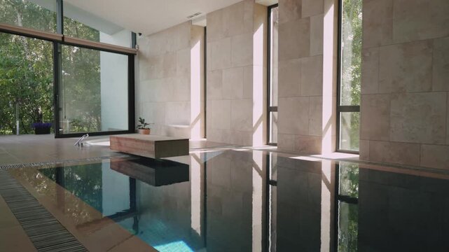 Smooth camera movement along indoor swimming pool in modern luxury interior, minimalist spa architecture with natural daylight, stone walls, large windows, calm water reflections, wellness lifestyle