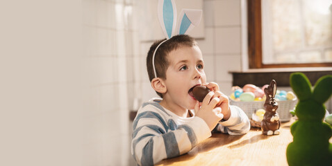 A boy dressed as a bunny eats a chocolate egg in the kitchen celebrating Easter.