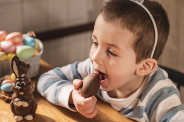 A boy dressed as a bunny eats a chocolate egg in the kitchen celebrating Easter.