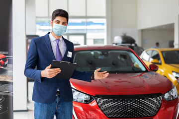 Arab man in formal outfit and protective face mask standing over luxury red car in big showroom, copy space. Male sales manager pointing at brand new automobile, holding chart with info about auto