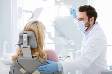 Obraz premium Male dentist in workwear, face mask and gloves having conversation with unrecognizable blonde woman sitting at dental chair, doctor greeting female patient, side view. Modern dentistry concept
