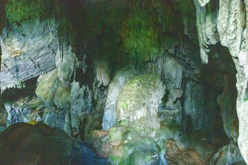 Organic limestone shapes and textured stalactites dominate the shadowy interior of Phu Kham Cave in Laos. Subtle green moss and mineral deposits highlight the rugged surfaces and natural patterns of