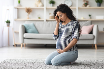 Curly long-haired millennial expecting lady feeling sick, sitting on floor in cozy living room and...