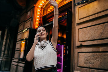 Young woman on phone raises her arm while smiling, combining dynamic body language with mobile communication — expressive digital presence in lively city atmosphere.