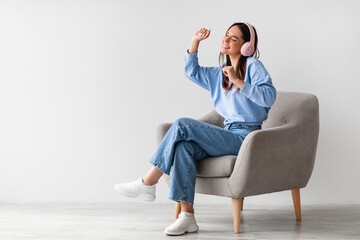 Happy millennial lady in wireless headphones relaxing in armchair and enjoying music against white...