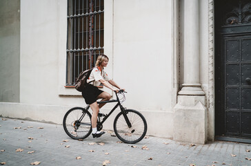 Asian woman rides bike past historical building, wearing floral shirt and backpack, symbolizing sustainable mobility, solo travel, and modern freedom with a minimalist urban lifestyle.