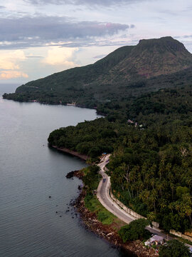 Aerial view of the rugged coastline meets the dense forest, framing the silhouette of a majestic mountain under a soft, diffused sky, Mambajao, Camiguin, Philippines.