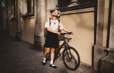 Young woman with headphones holds bicycle and smiles up at bright sky. Evokes hope, digital optimism, and freedom through tech and self-guided movement in urban surroundings.