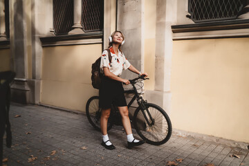 Smiling woman with bike and headphones stands on sunlit street, embracing moment with serenity and confidence. Urban mobility and personal tech use merge in stylish daily commute.