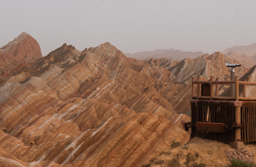 View of vibrant, striped sandstone formations rise majestically under a muted sky, with a wooden viewing platform, Zhangye, Gansu, China.